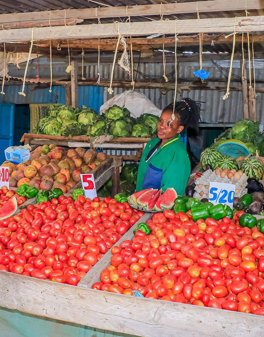 A grocer at her market stall with hand-written price cards among tomatoes and produce
